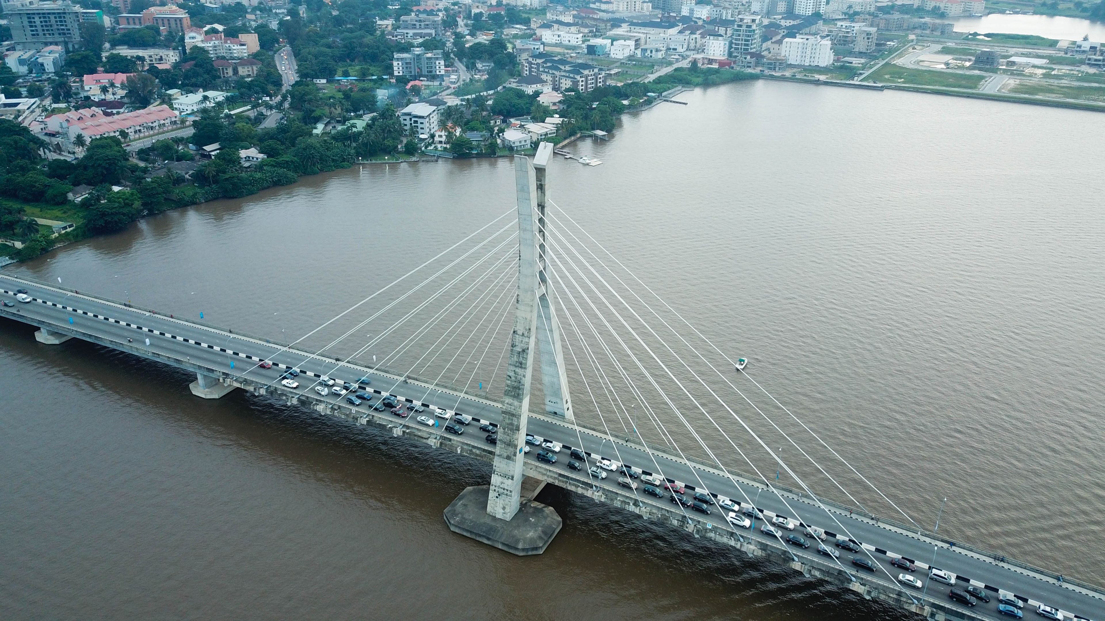 Eine Schrägseilbrücke über einem Fluss, im Hintergrund Autos, Gebäude und Grünflächen.