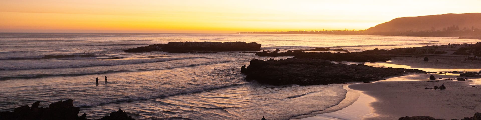 A beach at sunset with waves, rocks, and a distant city skyline.