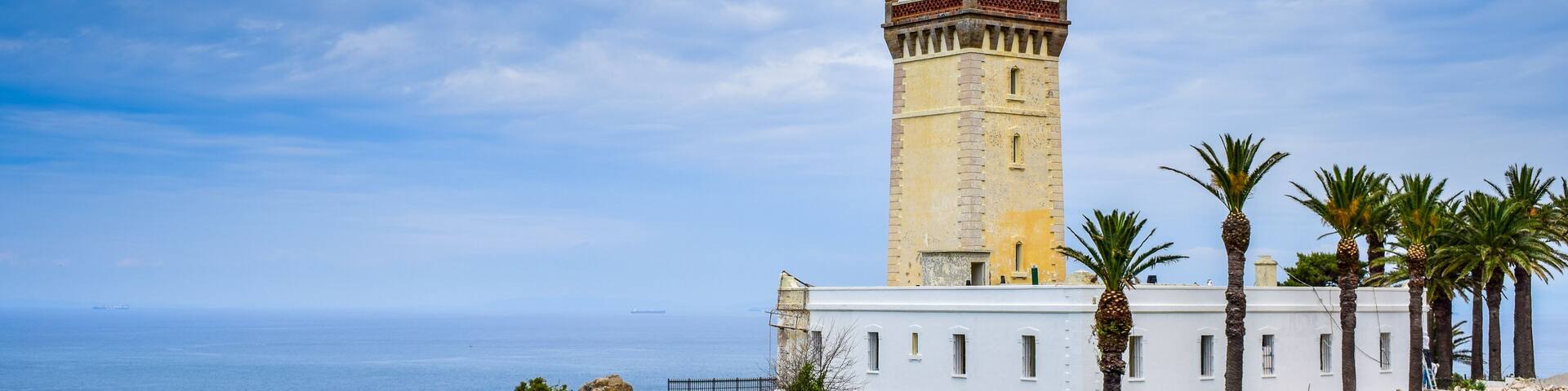 A coastal lighthouse with a white building nearby, palm trees, and the sea.