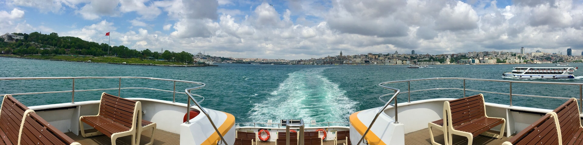 A boat deck with wooden benches, a wake in the water, and a cityscape in the background.