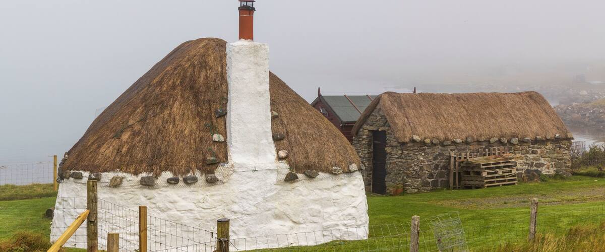 A thatched-roof house with a white exterior and a chimney, surrounded by a grassy field and a stone wall.