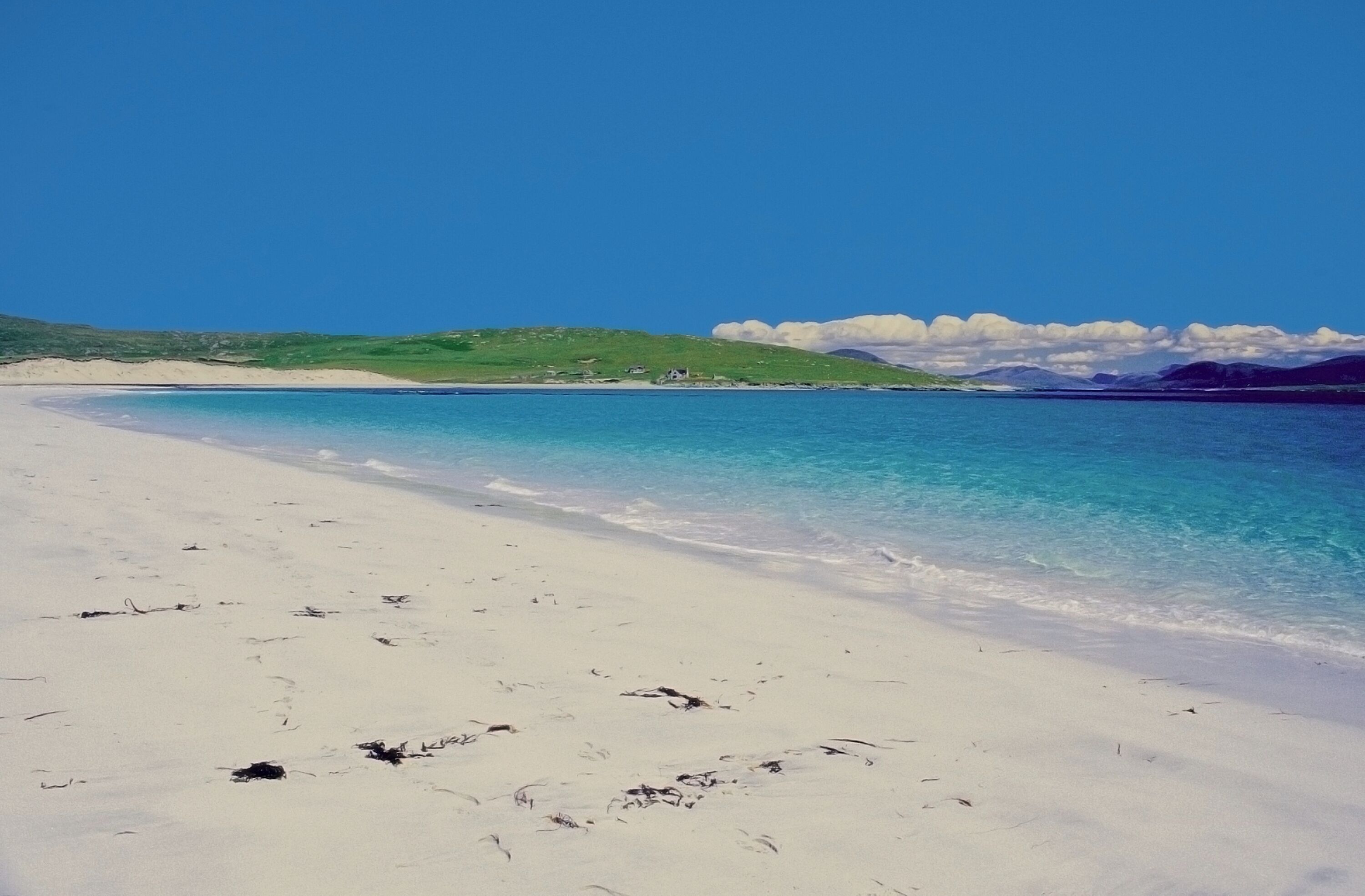 A beach with clear turquoise water, white sand, and green hills in the background.