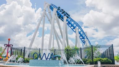 Uma montanha-russa com suportes brancos e uma pista azul, uma fonte de água e uma cerca com as letras "OWA" em primeiro plano.