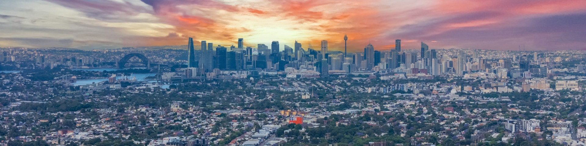 Une vue sur les toits d’une ville au coucher du soleil, avec de hauts gratte-ciel bien visibles et une zone résidentielle densément peuplée.