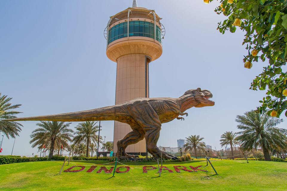 A dinosaur statue in a park with a tall tower in the background.