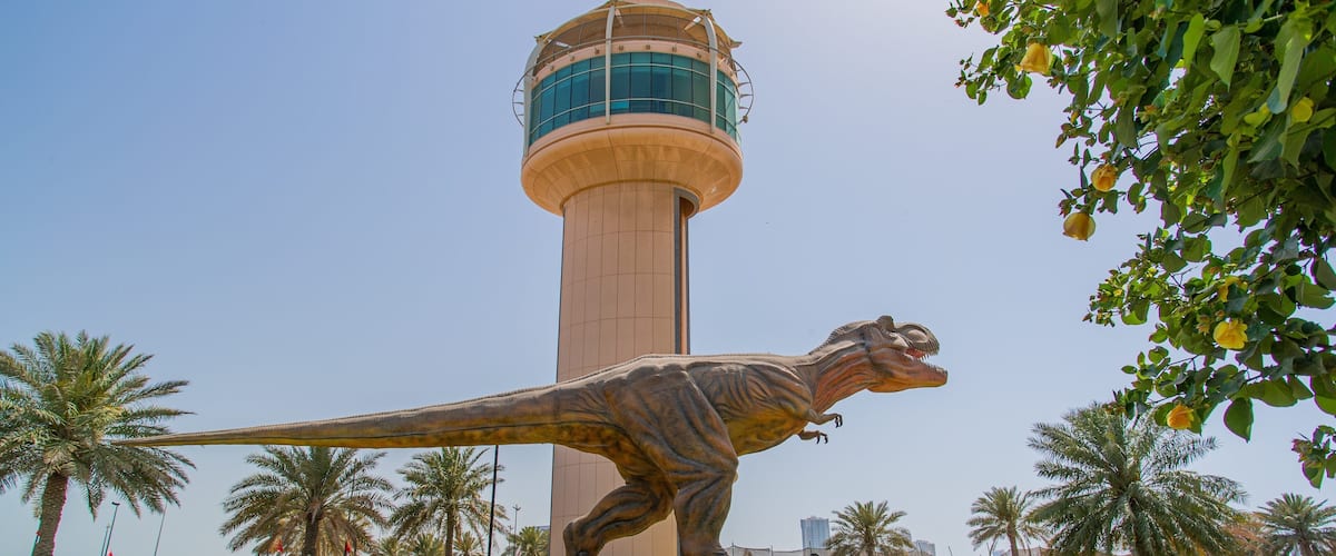 A dinosaur statue in a park with a tall tower in the background.