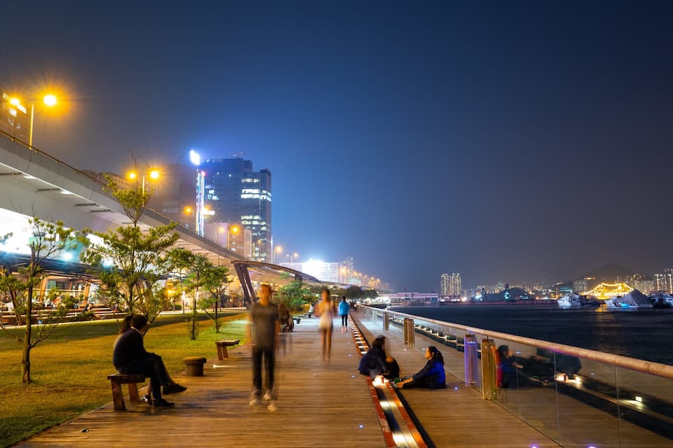 A riverside walkway at night with people walking and sitting.