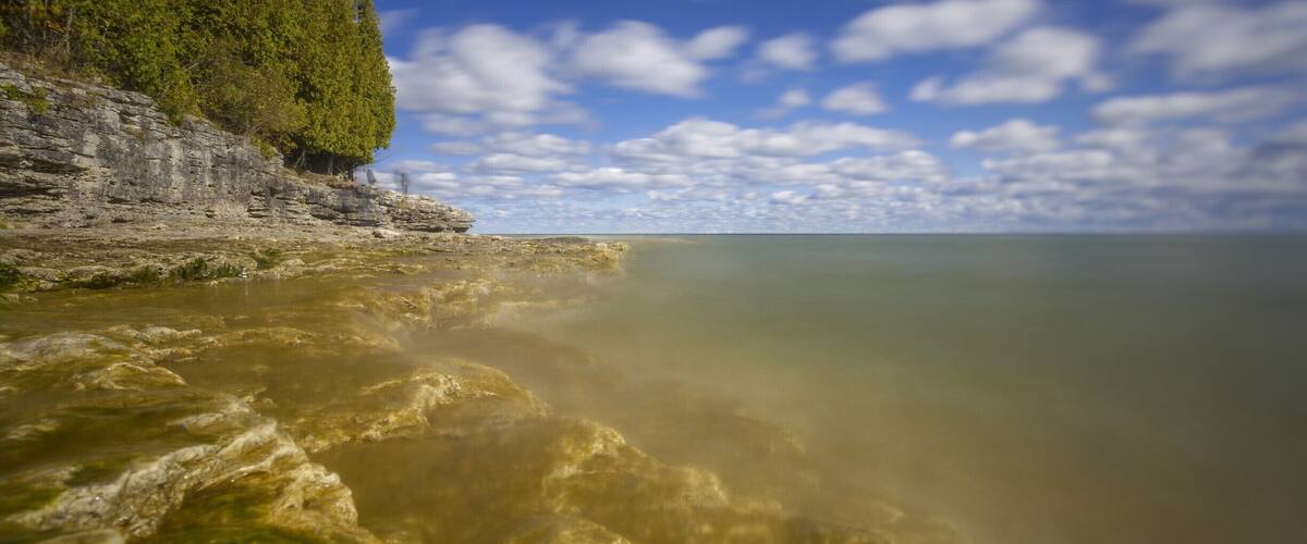 In Door county there is this small county park. It is very unassuming form the drive in. On its coast though lies stunning rock formations. The lake has eaten away at the soft rock and lowered the shelf so water is only a few inches deep.
I put on my super ND filter and just placed the camera on a small rock just above the water to take this. This actually looks away form the small caves that the park is known for.