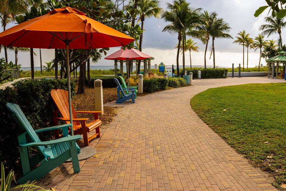 A paved walkway with green chairs and orange umbrellas, palm trees, and a gazebo.