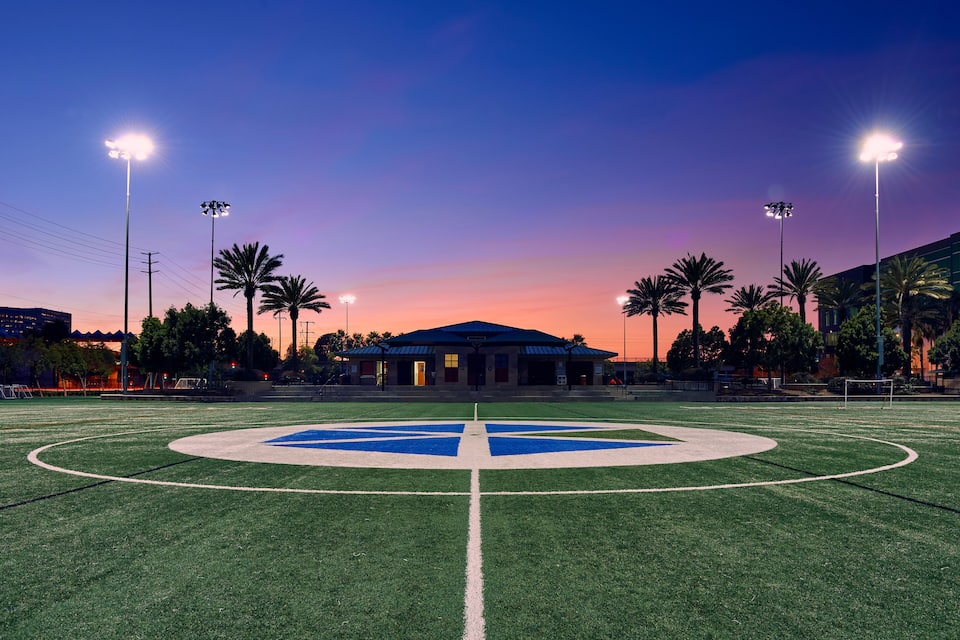 A football field at dusk with a large blue and white emblem in the center, surrounded by palm trees and a building with a blue roof.