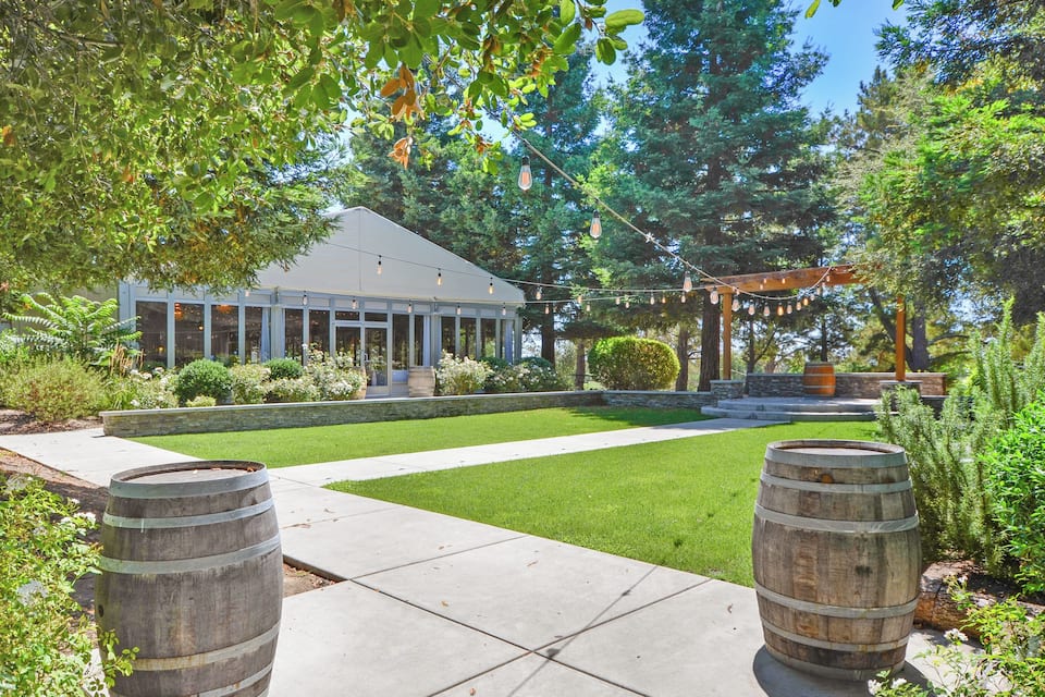 A well-maintained garden with a wooden pergola, a white building, and two wooden barrels.