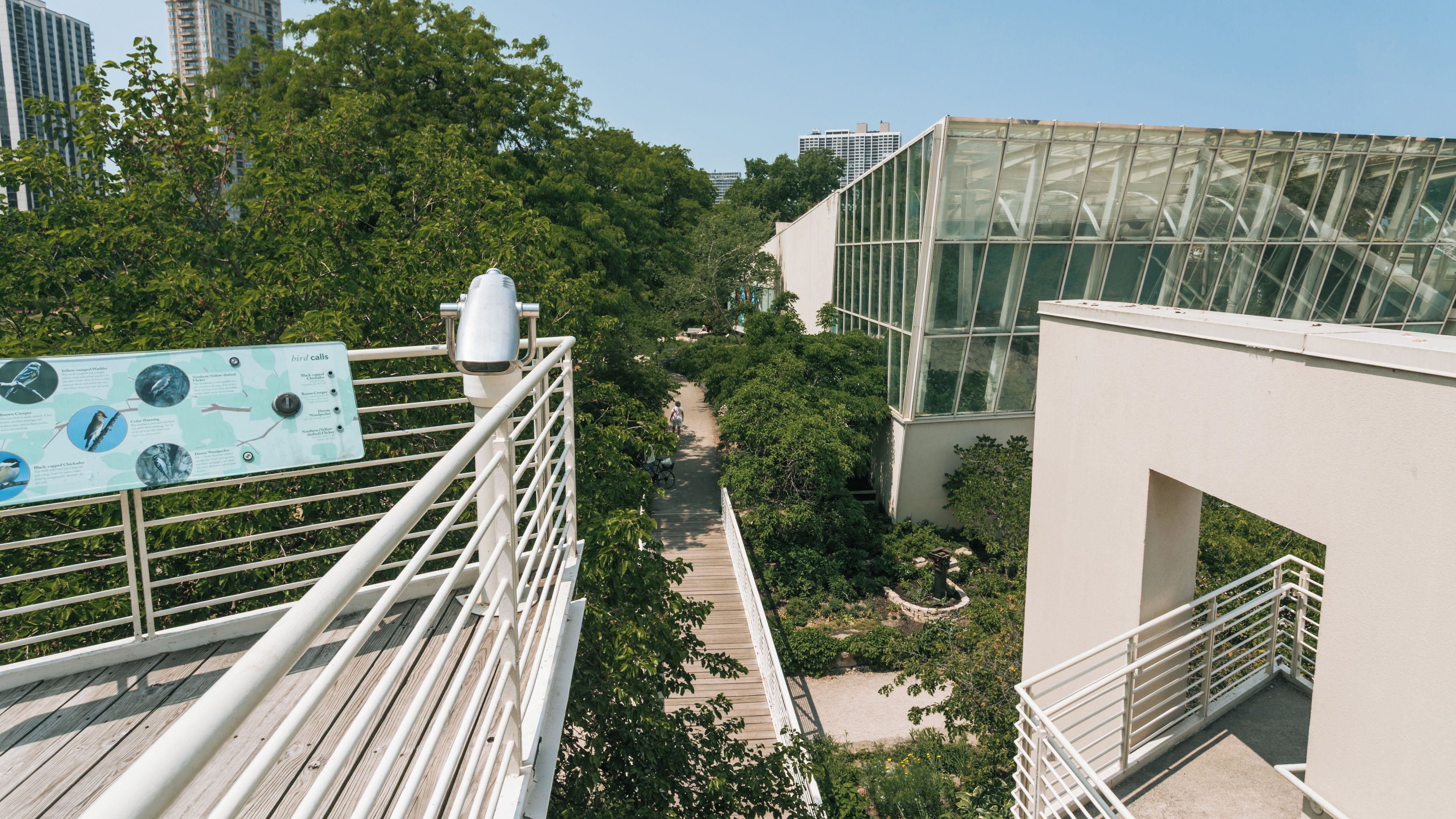 A pedestrian bridge with a glasshouse structure, surrounded by greenery and urban buildings.