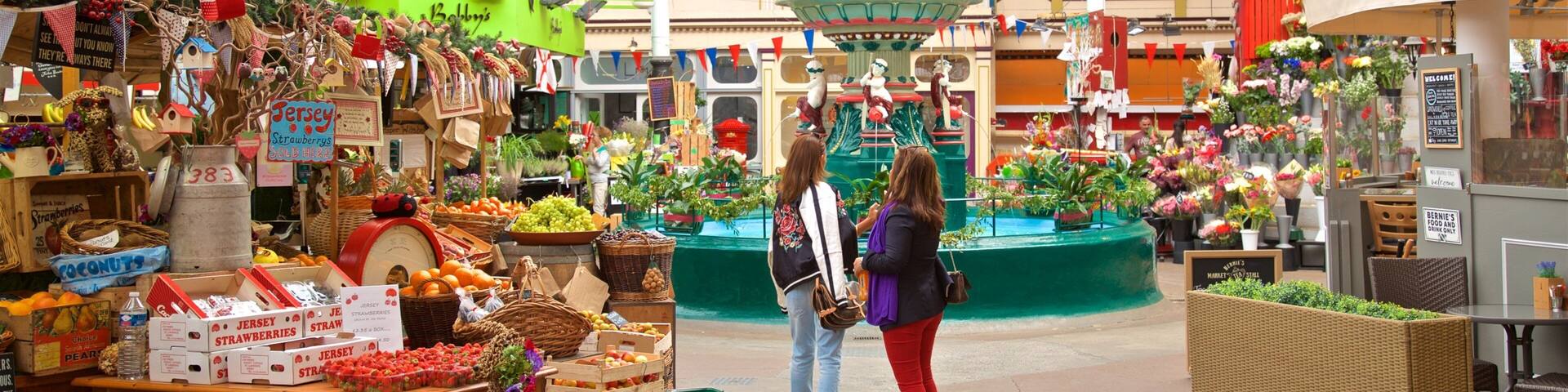 Un mercado interior con diversos puestos que ofrecen frutas, verduras y flores. Hay gente mirando los puestos.