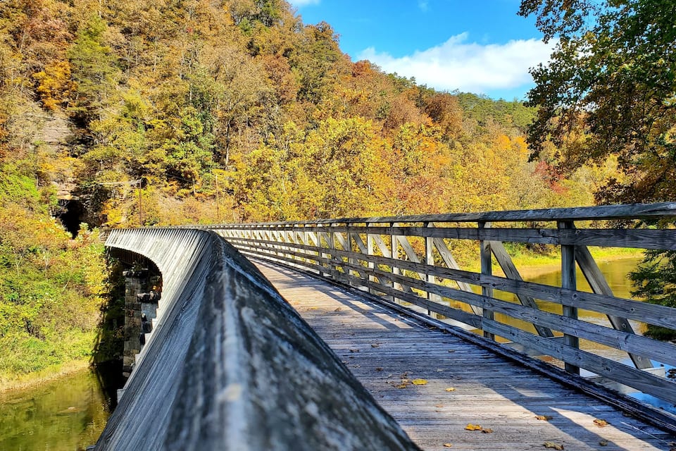A wooden bridge over a river, surrounded by autumn foliage and a forested hillside.