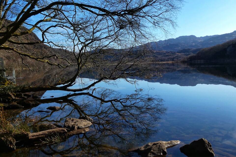 Um lago calmo com margens rochosas e árvores desnudas refletindo o céu.