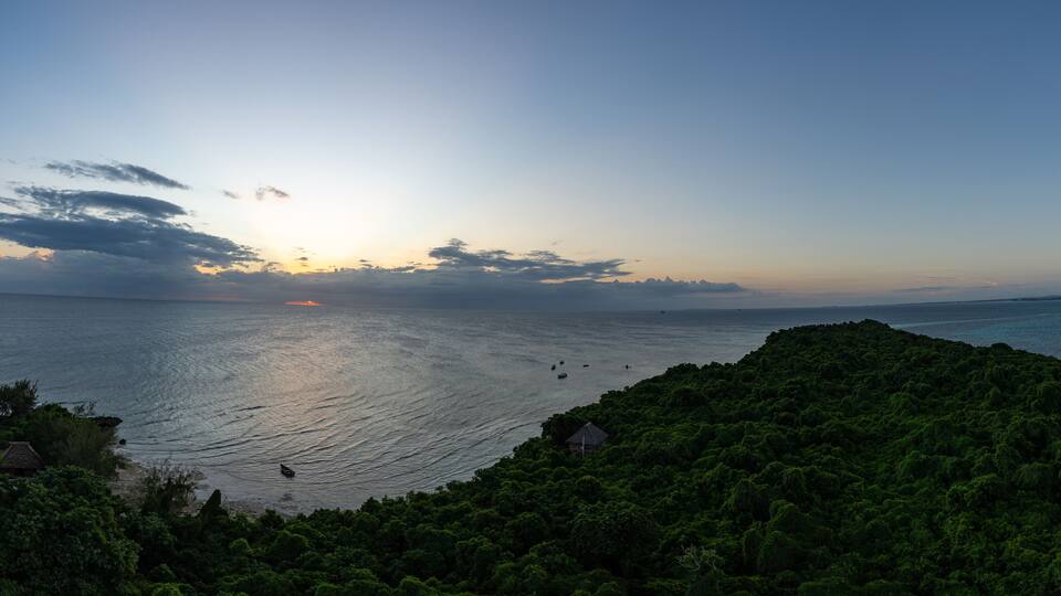 A coastal landscape with dense green vegetation, a calm sea, and a setting sun.