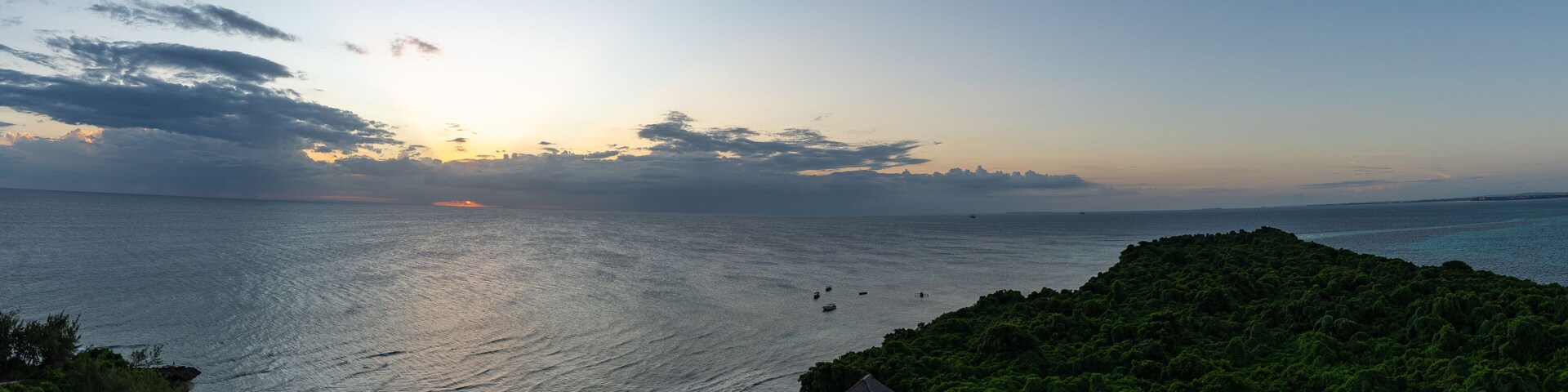 Sunset light fades over the Indian Ocean as it meets the lush green coastline of Chumbe Island, Zanzibar, with fishing boats scattered across the horizon.