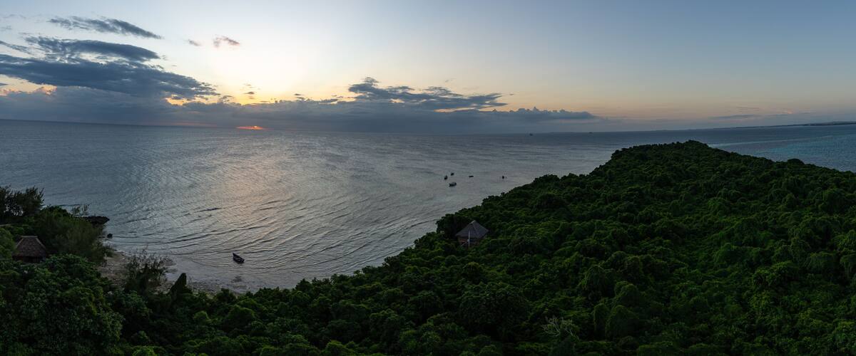 A coastal landscape with dense green vegetation, a calm sea, and a setting sun.