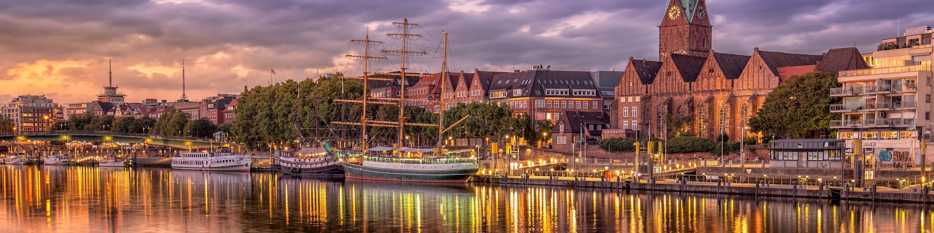 Paisagem urbana histórica à beira-mar, com uma imponente torre de igreja, um navio a vela tradicional e um rio calmo que reflete os edifícios.