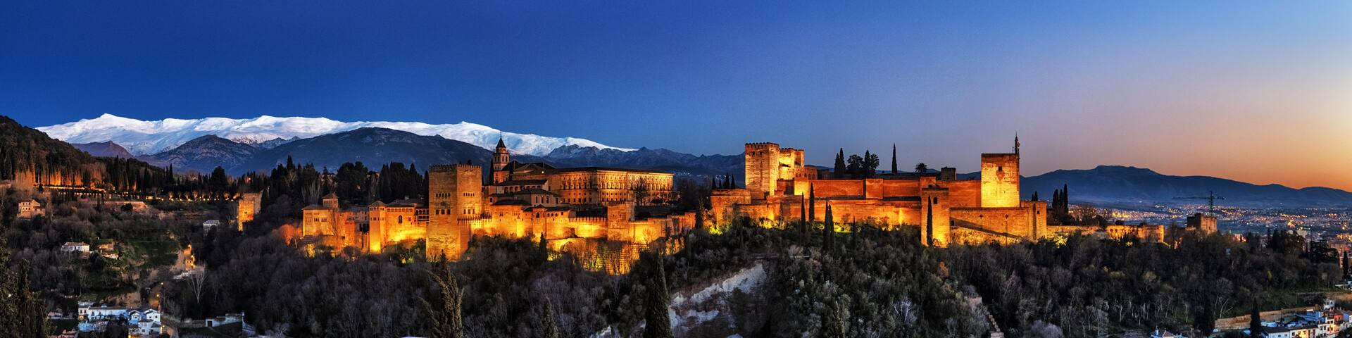 A historic castle complex with illuminated walls, a snow-capped mountain range in the background, and a cityscape at dusk.