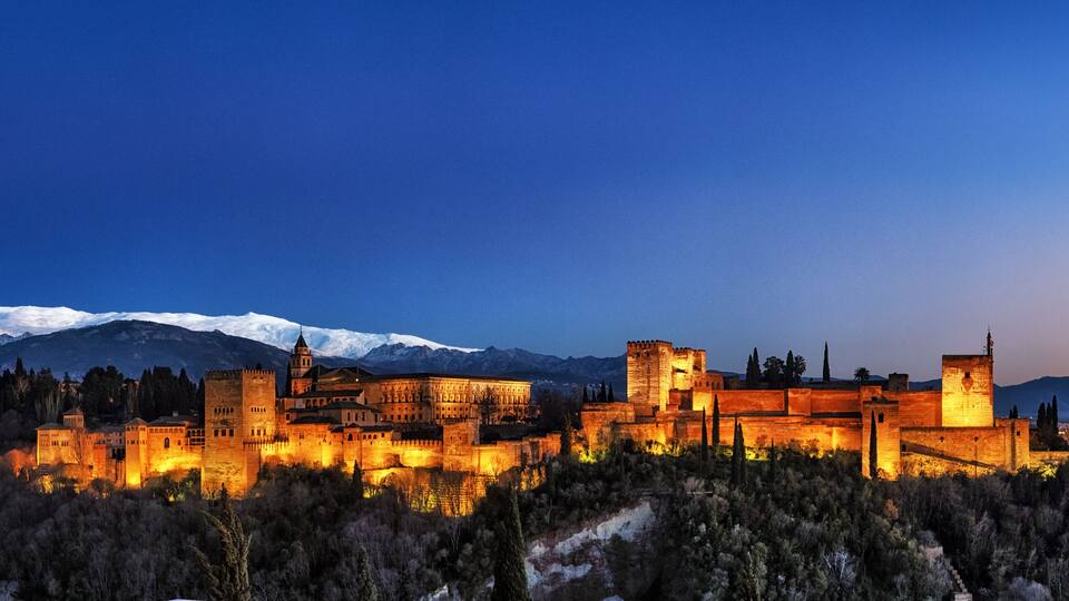 A historic castle complex with illuminated walls, a snow-capped mountain range in the background, and a cityscape at dusk.