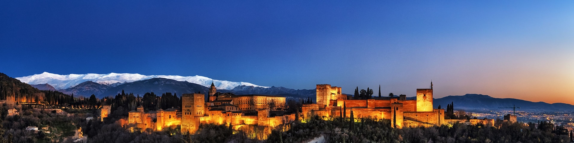 Um complexo de castelos históricos com muros iluminados, uma cordilheira nevada ao fundo e paisagem urbana ao entardecer.
