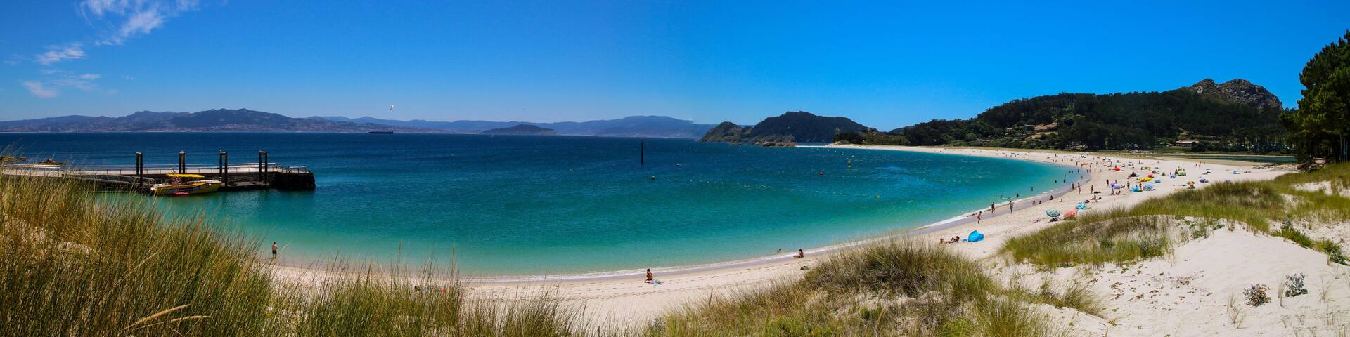 A beach with clear turquoise water, a pier, and distant mountains.