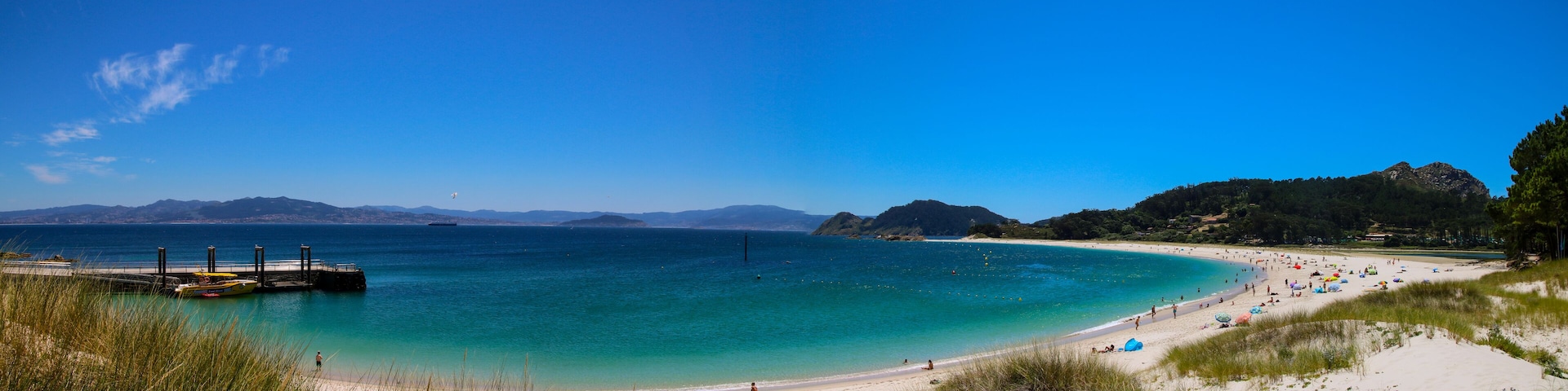 Ein Strand mit klarem türkisblauem Wasser, einer Mole und fernen Bergen.