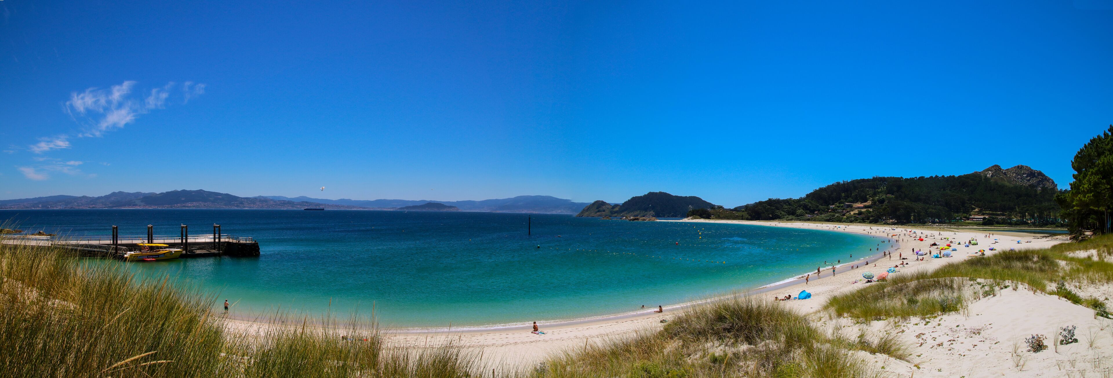 A beach with clear turquoise water, a pier, and distant mountains.