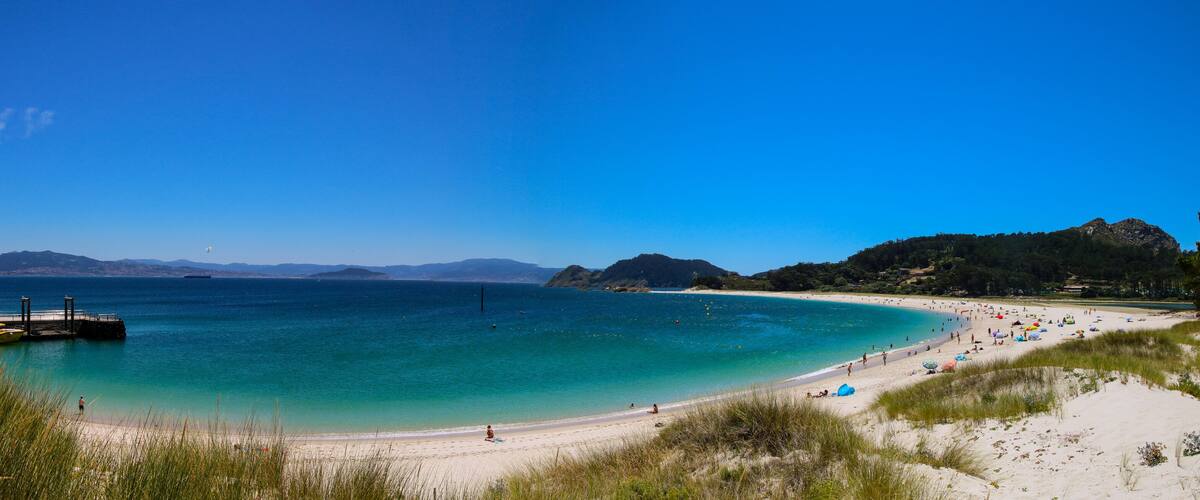 A beach with clear turquoise water, a pier, and distant mountains.