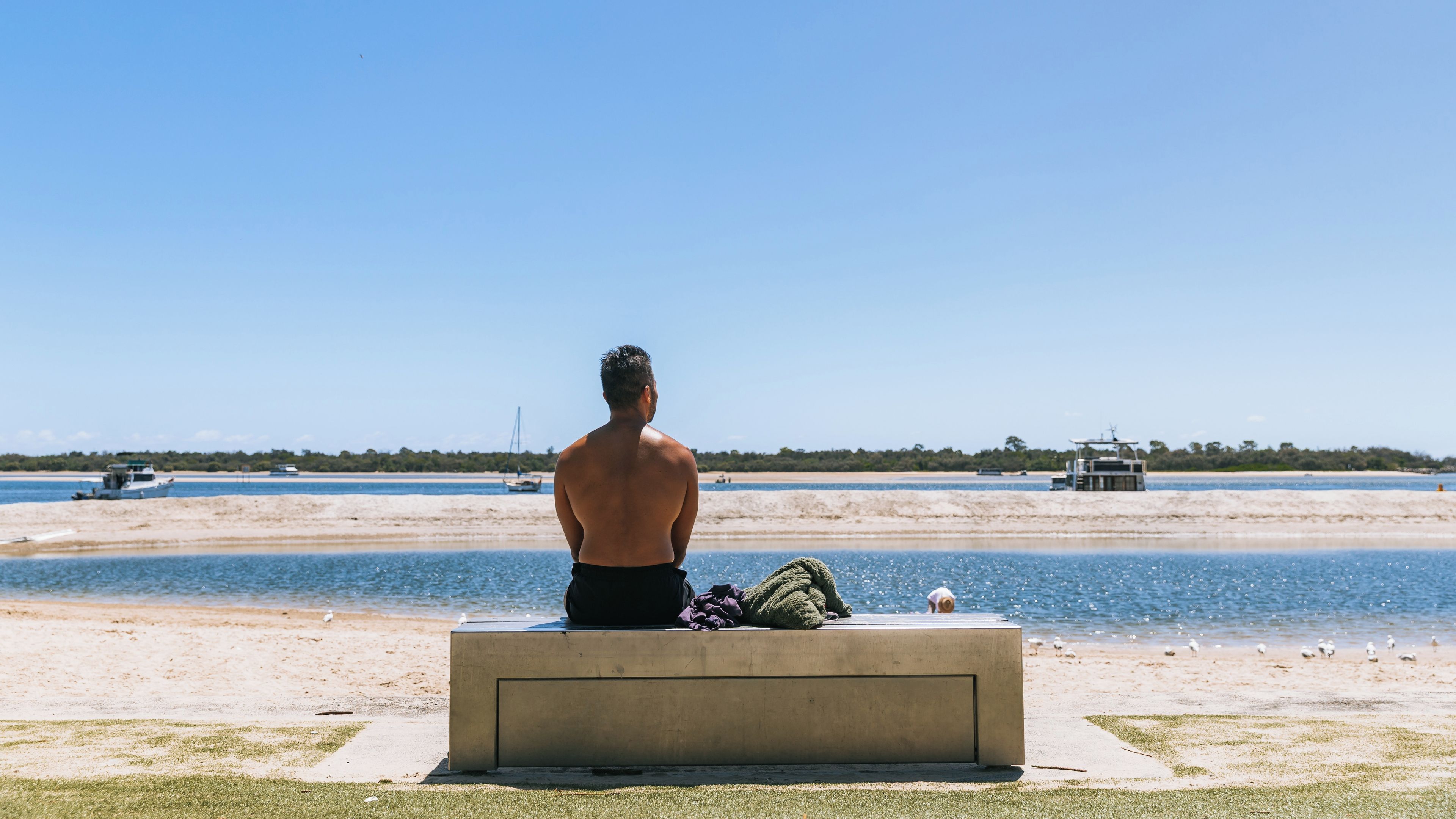A man sitting on a bench overlooking a beach with boats and a lighthouse in the distance.