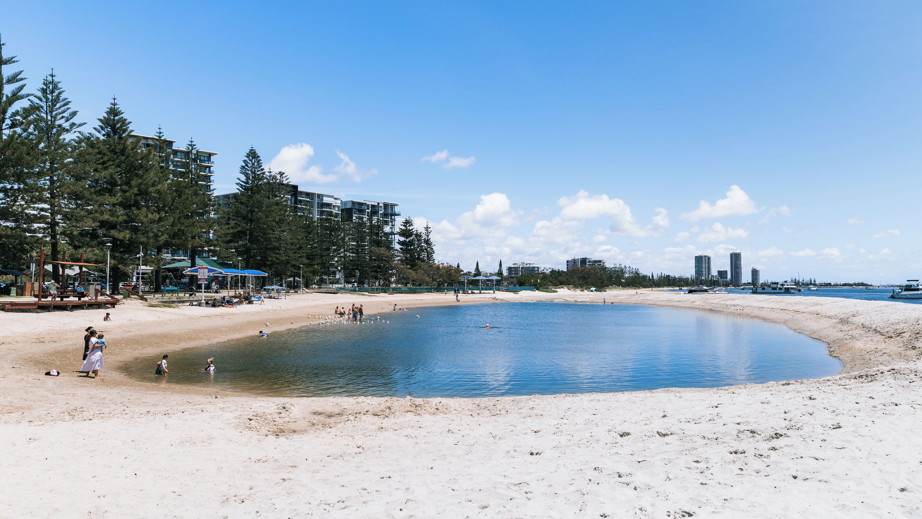 A beach with a calm water body, sandy shore, and buildings in the background.