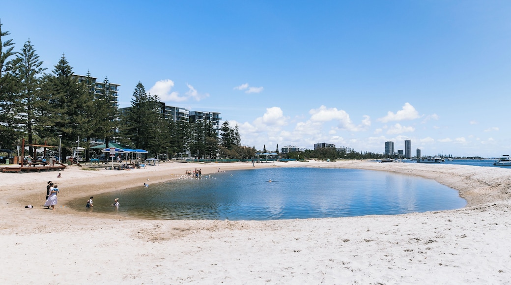 A beach with a calm water body, sandy shore, and buildings in the background.