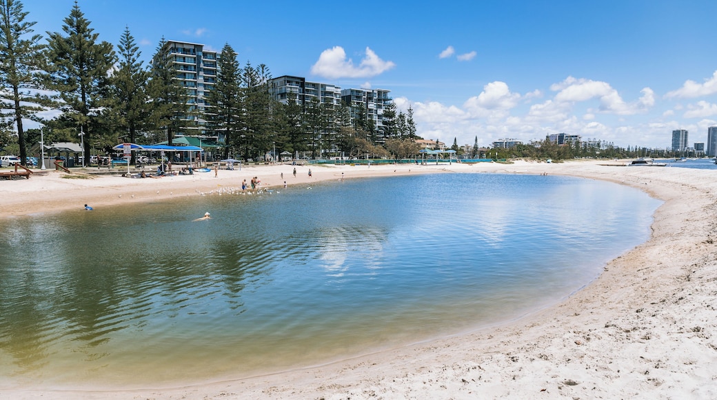 A sandy beach with clear water, a few people, and tall buildings in the background.