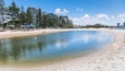 A sandy beach with clear water, a few people, and tall buildings in the background.