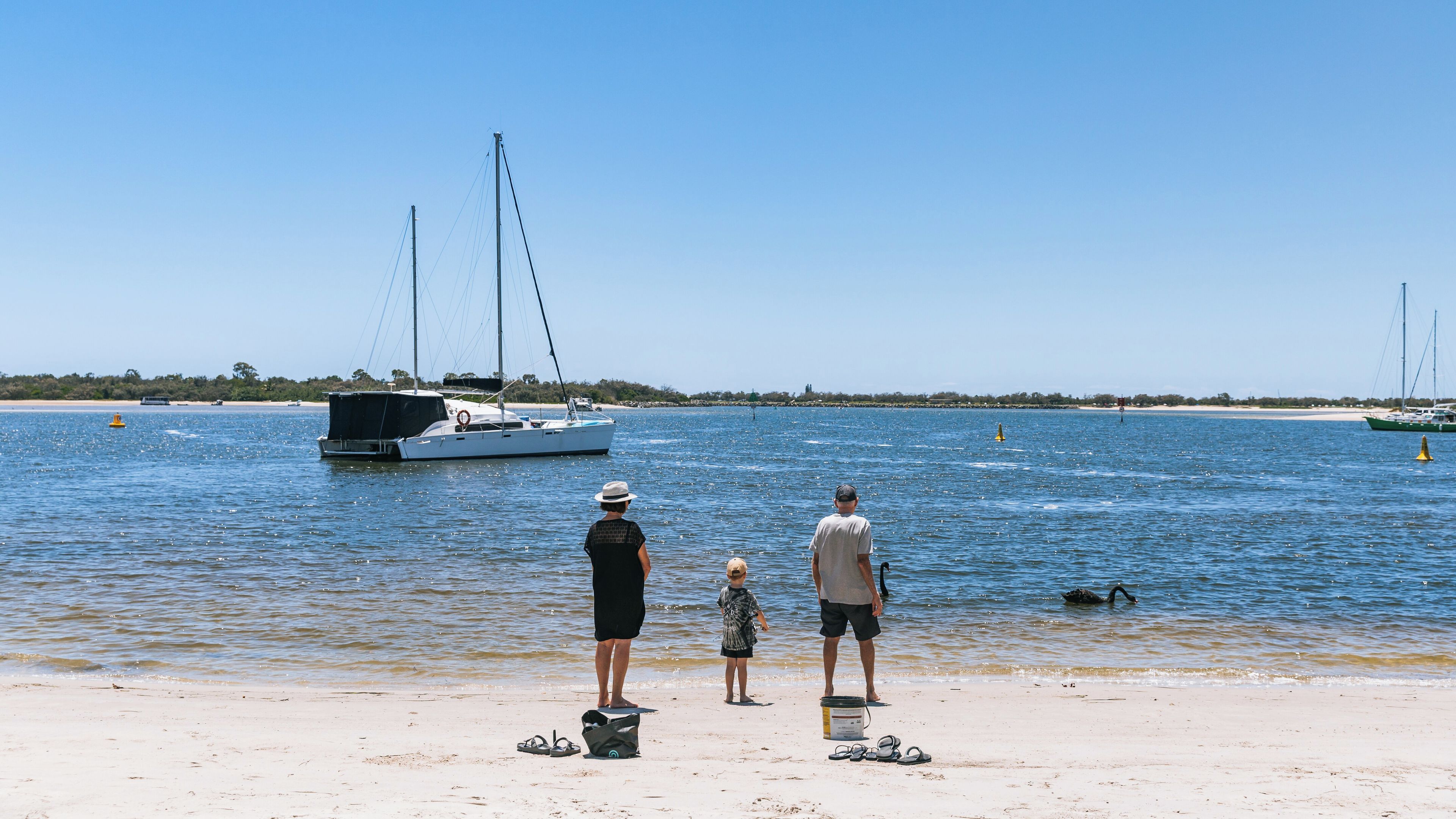 A beach with a sailboat in the water, a lifebuoy on the sand, and a child standing between two adults.