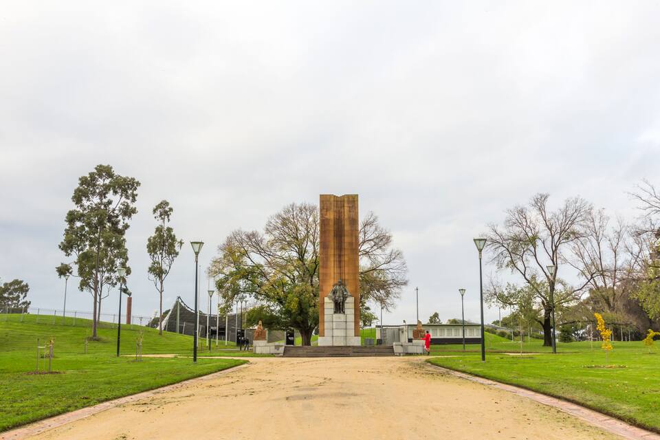 Een monument in een park, met een pad van aarde dat erheen leidt, omringd door bomen en lantaarnpalen.