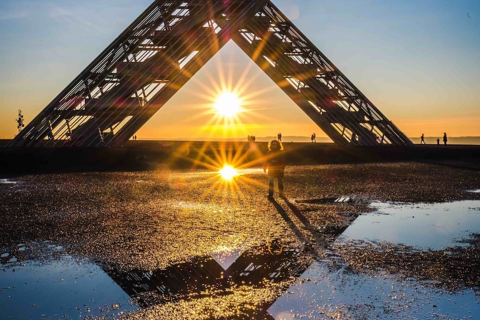 A large metal structure with a star-like shape, reflecting the sunset over a body of water.