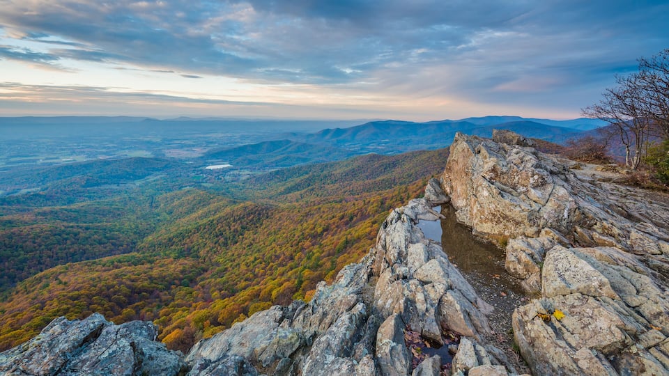A rocky overlook with a panoramic view of a forested valley and distant mountains under a partly cloudy sky.