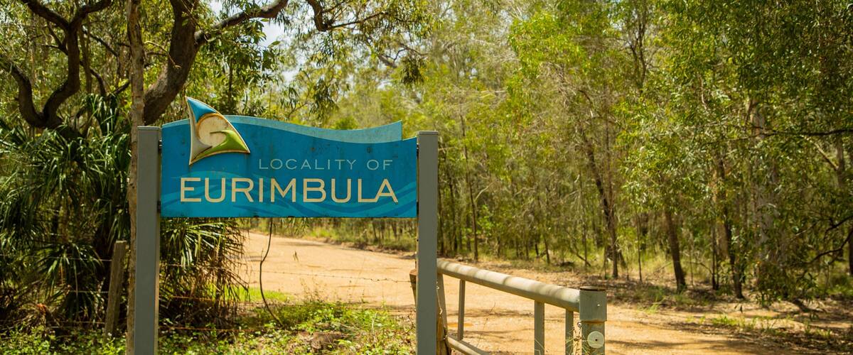 Un panneau indiquant l’entrée de la localité d’Eurimblia, avec une clôture en bois et des arbres en arrière-plan.