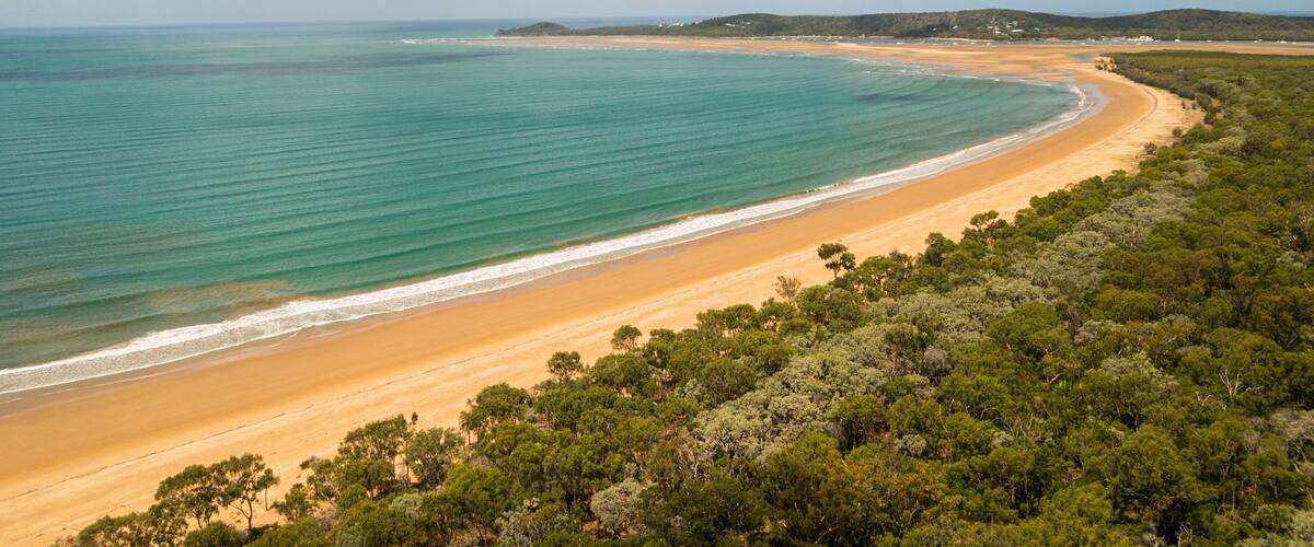 A coastal landscape with a sandy beach, turquoise waters, and dense green vegetation.