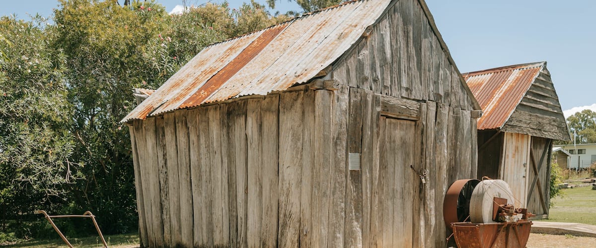 A rustic wooden shed with a corrugated metal roof, a wheelbarrow, and a plow in front.