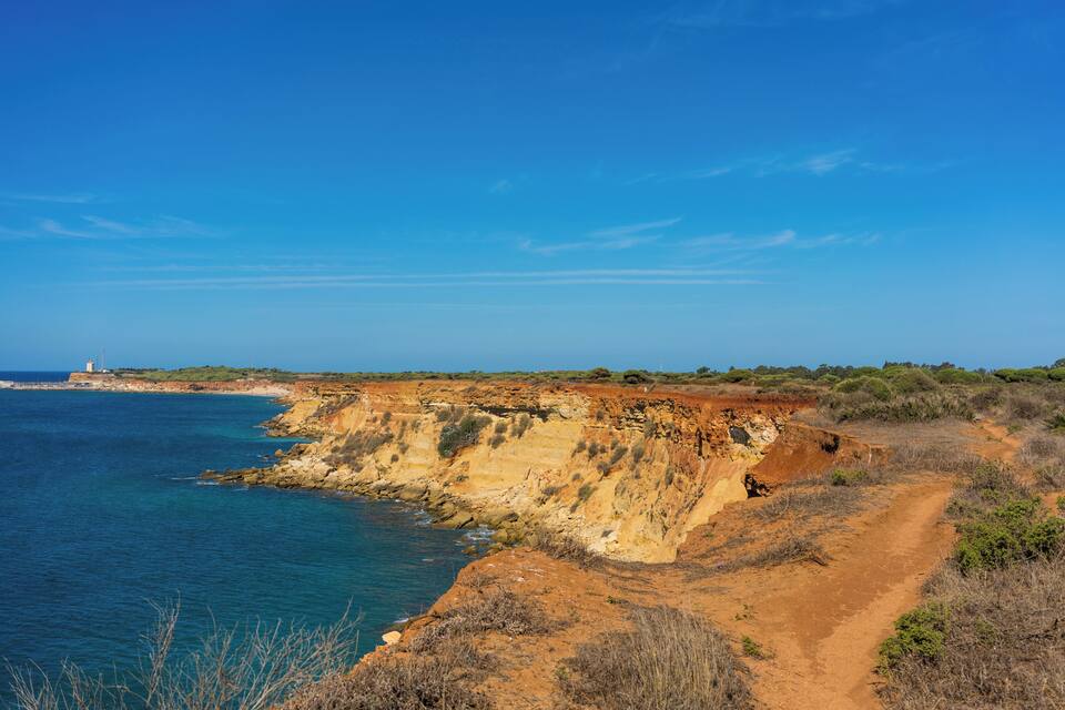 A coastal landscape with cliffs, a lighthouse, and a dirt path along the shoreline.