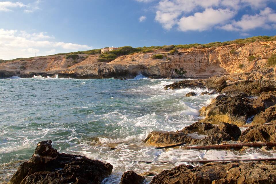 Une côte rocheuse où les vagues s’écrasent sur le rivage, une falaise à la végétation clairsemée et un bâtiment situé sur le versant de la colline.