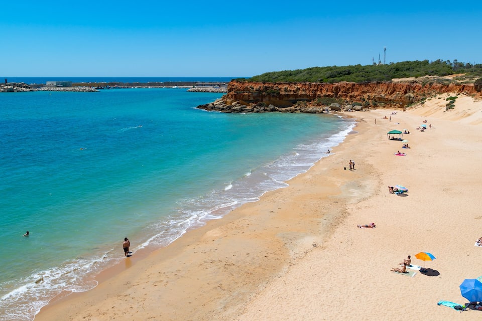 A sandy beach with clear turquoise water, a rocky cliff, and a few people enjoying the sun.