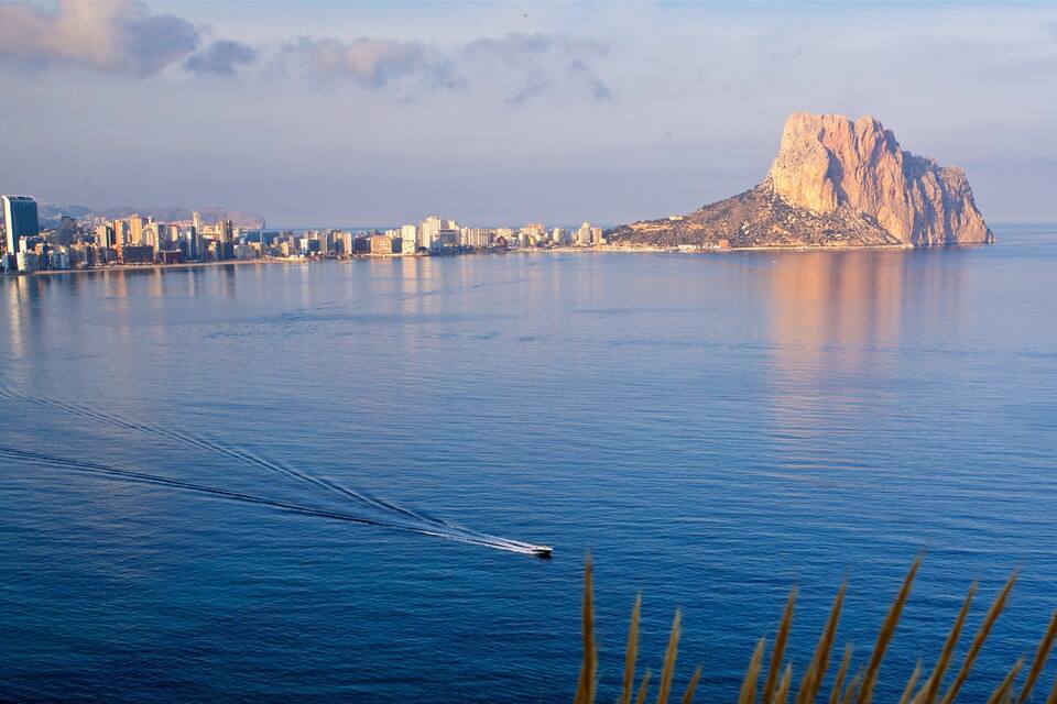 A coastal cityscape with a prominent rocky hill and calm blue waters.