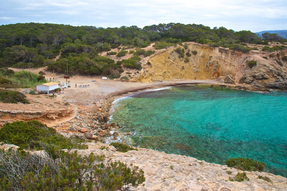A coastal landscape with a clear turquoise sea, rocky cliffs, and a small building on the beach.