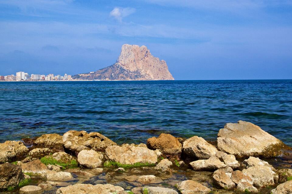Una costa rocosa con agua cristalina, una montaña distante y el perfil de una ciudad.