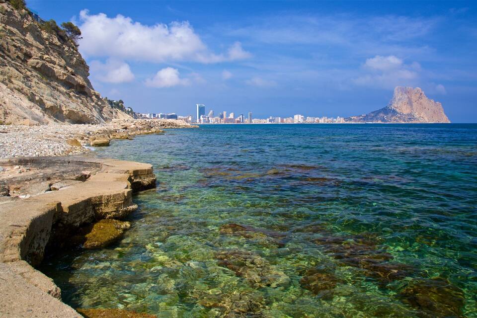A coastal landscape with clear waters, rocky shore, and a distant city skyline.