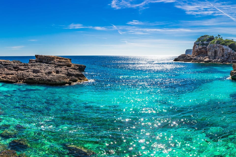A coastal landscape with clear turquoise waters, rocky formations, and a cliff with vegetation.